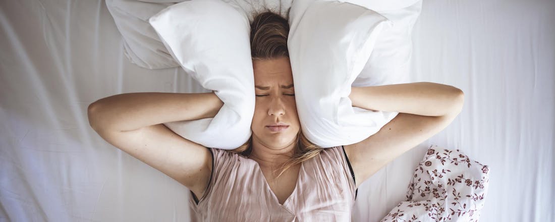 Vrouw in bed met kussen over haar oren wegens geluidsoverlast Wat kun je doen bij overlast van de buren?