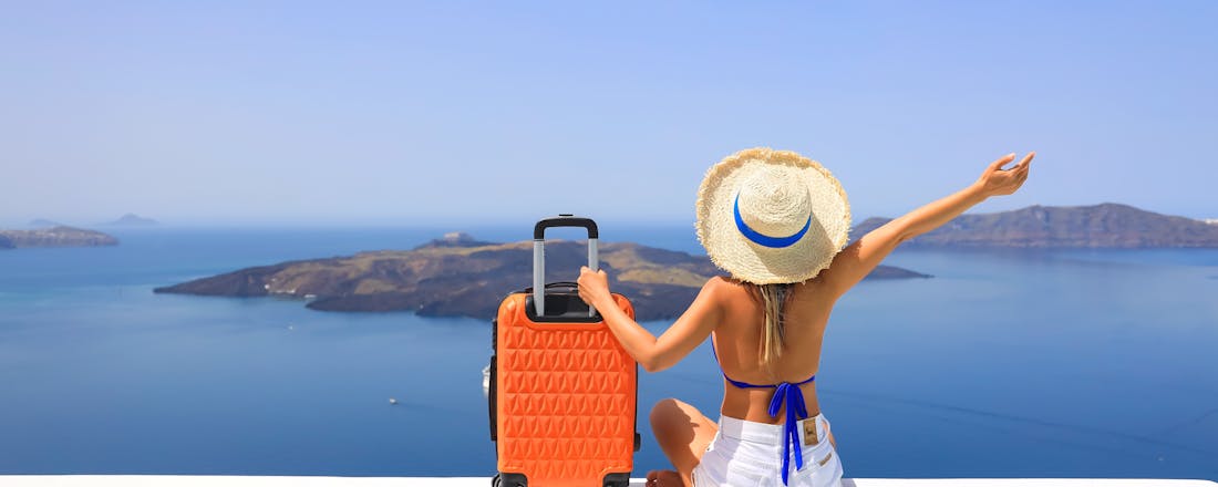 vrouw met koffer op de rand van een stenen muur kijkend naar de zee Acht tips voor een zorgeloze vakantie